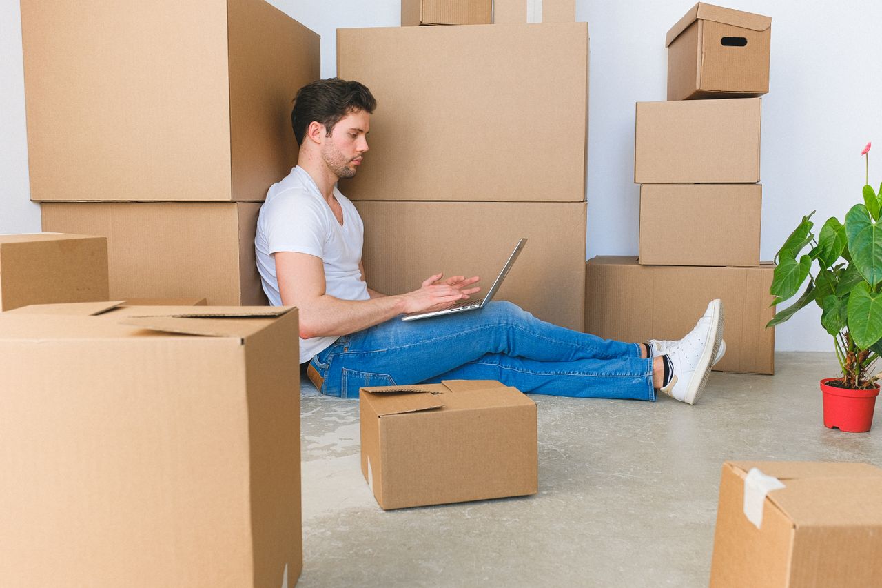 Man sitting on floor with laptop surrounded by stacked cardboard moving boxes
