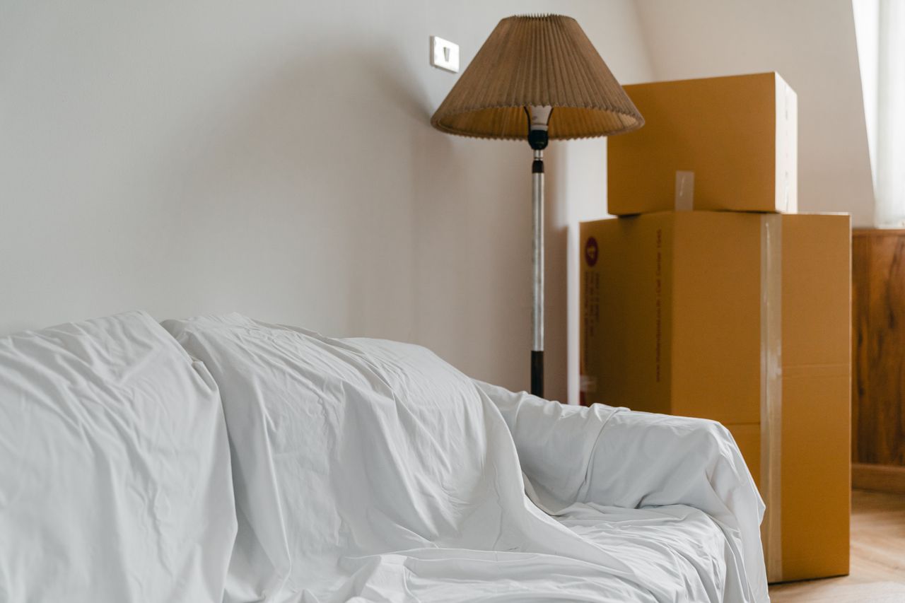 Sofa covered with a white dust sheet next to stacked cardboard moving boxes and a floor lamp